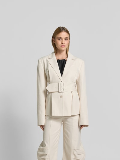Frauenmodel trägt Blazer aus Leinen-Mix mit Taillen-Gürtel von ROTATE in Creme mit Reverskragen, Knopfleiste und Brusttasche, taillierte Passform, Ganzkörperansicht von vorne.