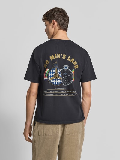 Männermodel trägt Regular Fit T-Shirt aus Baumwolle von RHUDE mit Label-Print auf dunklem Stoff, Rückenansicht, Ganzkörperansicht von hinten.