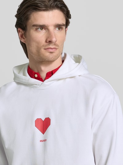 Männermodel trägt Regular Fit Hoodie aus reiner Baumwolle Modell 'DALENTINOS' von HUGO, Weiß mit rotem Motiv-Print, Nahaufnahme Oberkörper.