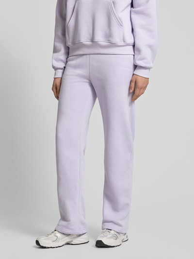 Kobiece model prezentuje Sweatpants mit elastischem Bund Modell 'Iconic' od Gina Tricot, bawełniany miks, krój relaxed fit, kolor jasnofioletowy, widoczny elastyczny pas i tunel na sznurek, długość nogawki 80 cm w rozmiarze S, zdjęcie całej sylwetki z przodu.