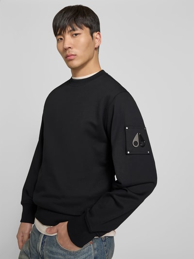 Männermodel trägt Sweatshirt mit Rundhalsausschnitt von Moose Knuckles in Schwarz, Reine Baumwolle, Regular-Fit, Logo-Detail am Ärmel, elastische Abschlüsse, Rückenlänge bei Größe M 69 cm, Ganzkörperansicht von vorne.