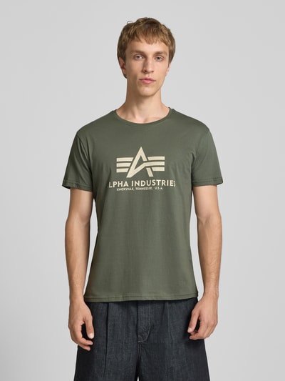 Mannelijk model draagt T-shirt met Logo en Rundhalsausschnitt van Alpha Industries in olijfgroen, puur katoen, regular fit, logoprint, ronde hals, ruglengte bij maat M 68 cm. Ganzkörperansicht van voren.