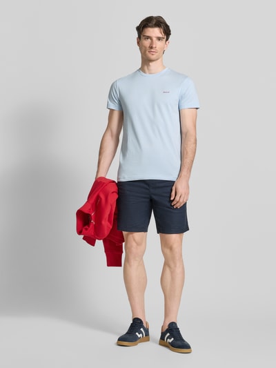 Męski model nosi t-shirt z detalem z logo od Gant w jasnoniebieskim kolorze, bawełna, prosty krój, okrągły dekolt, długość tyłu w rozmiarze M: 70 cm. Ganzkörperansicht von vorne.