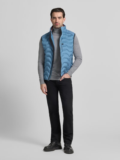 Männermodel trägt Regular Fit Steppweste mit Kapuze im Kragen von Tom Tailor in Hellblau, kombiniert mit grauem Rollkragenpullover, Ganzkörperansicht von vorne. Unifarbene Optik, reflektierende Details, Label Patch, Reißverschluss, seitliche Taschen sowie Innentasche sichtbar.