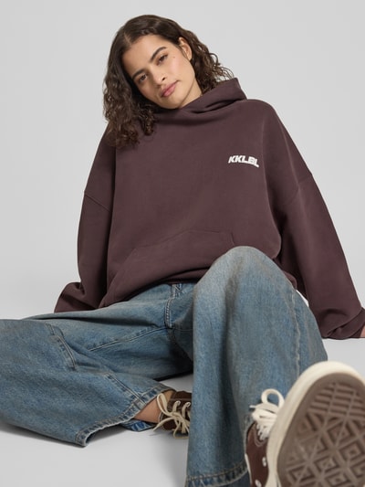 Frauenmodel trägt Oversized Hoodie mit Label-Prints und Känguru-Tasche von Karo Kauer in Dunkelbraun, kombiniert mit weiter Jeans, reine Baumwolle, Nahaufnahme Oberkörper.