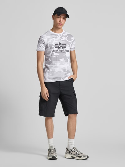 Męski model nosi T-Shirt mit Label-Print von Alpha Industries w kolorach szaro-białych, bawełna, prosty krój, okrągły dekolt, długość tyłu w rozmiarze M: 70 cm, Ganzkörperansicht von vorne.