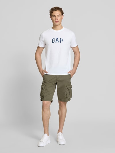 Männermodel trägt T-Shirt mit Logo-Patch und Rundhalsausschnitt von GAP aus reiner Baumwolle, Straight Fit, weiß, Ganzkörperansicht von vorne.