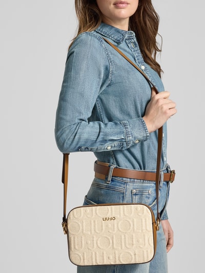Frauenmodel trägt Camera Case mit Logo-Detail Modell 'Manhattan' von Liu Jo Jeans, beigefarbene Textur, rechteckige Form, Nahaufnahme Oberkörper. Hauptfach mit Reißverschluss, schmaler Schulterriemen, Maße 23x16x8 cm, Riemenlänge 130 cm.