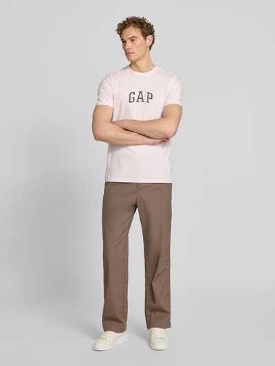 Männermodel trägt T-Shirt mit Logo-Patch und Rundhalsausschnitt von GAP aus reiner Baumwolle, Straight Fit, hellrosa Farbe, Ganzkörperansicht von vorne.