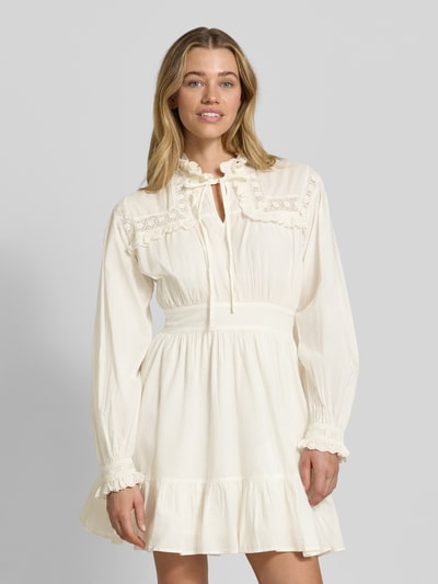Frauenmodel trägt knielanges Kleid mit Rüschenbesatz Modell 'Gilara' von mbyM aus Reine Baumwolle, Tailliert, Langärmelig, Stehkragen, Schlüssellochausschnitt, eingefasste Ärmelabschlüsse, Farbe Creme, Ganzkörperansicht von vorne.