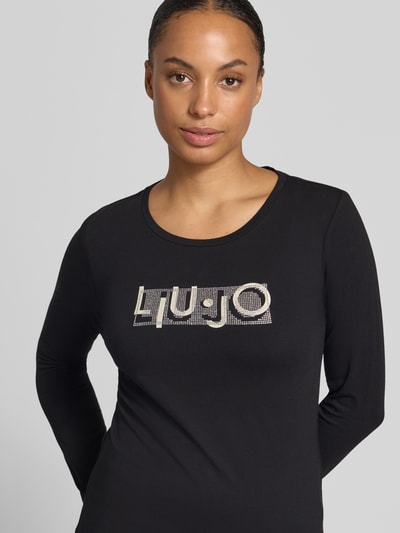 Frauenmodel trägt Langarmshirt aus Baumwoll-Mix von Liu Jo Sport in Schwarz mit Rundhalsausschnitt, Label-Print und Ziersteinen, Nahaufnahme Oberkörper.