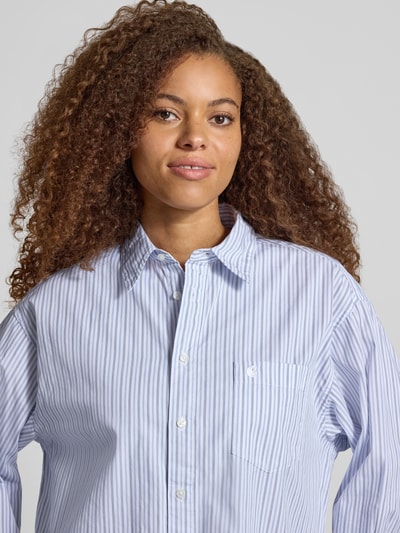 Vrouwenmodel draagt Relaxed Fit blouse met labelstitching Model 'Keene' van Carhartt Work In Progress, puur katoen, overhemdkraag, knoopsluiting, borstzak, lichtblauw-wit gestreept, textuur glad, Nahaufnahme Oberkörper. Ruglengte bij maat S: 78 cm, mouwlengte bij maat S: 56 cm.