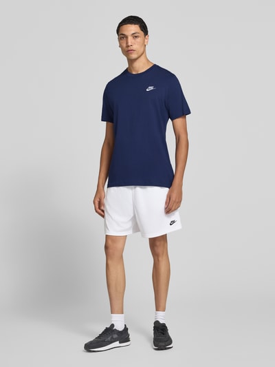 Männermodel trägt Regular Fit T-Shirt aus reiner Baumwolle von Nike in Dunkelblau, weiße Shorts, weiße Wand, Ganzkörperansicht von vorne. Herren T-Shirt von Nike