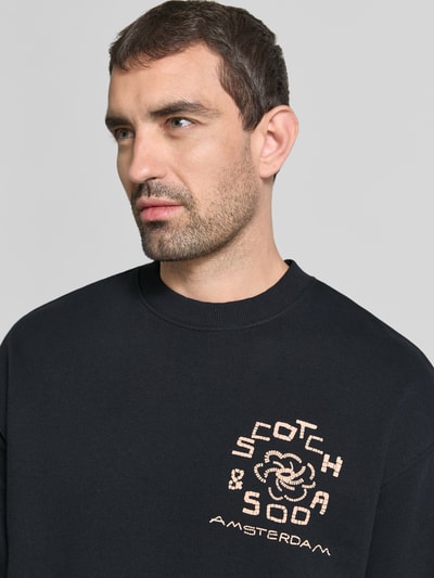 Mannelijk model draagt Relaxed Fit Pullover mit Logo-Print van Scotch & Soda in zwart met logoprint, puur katoen, ronde hals, elastische boordjes, Nahaufnahme bovenlichaam.