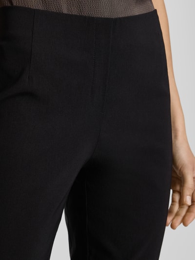 Kobieta nosi spodnie materiałowe z zakładkami w pasie, model 'INA' od Stehmann, jednokolorowy design, detal, zbliżenie bioder. Szerokość w pasie przy rozmiarze 36: 38 cm, długość wewnętrzna nogawki: 99 cm.