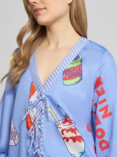Modelka nosi Bluse mit Schnürung und V-Ausschnitt von Milano Italy w kolorze jasnoniebieskim z kolorowym nadrukiem, luźny krój, czysty bawełna, dekolt w serek, sznurowanie, zapięcie na guziki, balonowe rękawy, długość pleców w rozmiarze 36: 57 cm, długość rękawa w rozmiarze 36: 62 cm; zbliżenie na górną część ciała, widok z przodu.