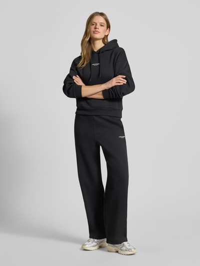Frauenmodel trägt High Waist Sweatpants mit elastischem Bund und logo von JJXX, mieszanka bawełny, szerokie nogawki, długość wewnętrzna 74 cm, czarne, gładka tekstura, całkowity widok z przodu.