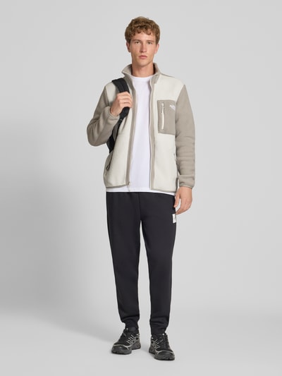 Męski model nosi kurtkę z polaru z kieszeniami zapinanymi na zamek błyskawiczny, model 'YUMIORI' od The North Face, luźny krój, stójka, kolor jasnoszary i beżowy, z elastycznymi wykończeniami, długość tyłu 71 cm, długość rękawów 71 cm, widok całej sylwetki z przodu.