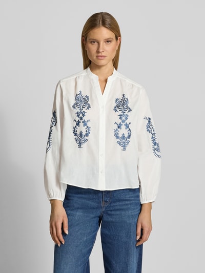 Frauenmodel trägt Regular Fit Bluse mit floralem Stitching Modell 'Februar' von More & More aus Reiner Baumwolle, Weiß mit blauen Details, Langärmelig, elastische Ärmelabschlüsse, Tunika-Ausschnitt, Knopfleiste, Rückenlänge 60 cm, Ärmellänge 60 cm, Ganzkörperansicht von vorne.