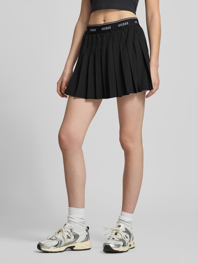 Frauenmodel trägt Faltenrock mit elastischem Bund Modell 'SELENE' von Guess Activewear, schwarzer Minirock in A-Linie, Material-Mix mit Stretch-Anteil, Bundweite 35 cm, Gesamtlänge 36 cm, Ganzkörperansicht von vorne.