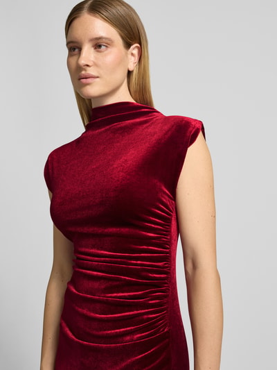 Frauenmodel trägt Cocktailkleid mit seitlicher Raffung von APRICOT in unifarbenem, samtigem Rot, hochgeschlossen, ärmellos, Nahaufnahme Oberkörper.