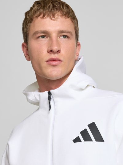 Männermodel trägt Sweatjacke mit Kapuze von adidas Sportswear in Weiß mit schwarzem Label-Print, Material-Mix, Nahaufnahme Oberkörper.
