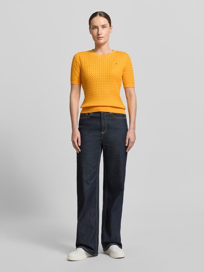 Frauenmodel trägt Slim Fit T-Shirt aus reiner Baumwolle von Tommy Hilfiger in Optik: Unifarben mit Zopfmuster, Logo-Detail, Rundhals, Kurzarm, gerippte Abschlüsse, Strick aus Reine Baumwolle, kräftiges Gelb, kombiniert mit blauer Hose, Strukturmuster sichtbar, Ganzkörperansicht von vorne.