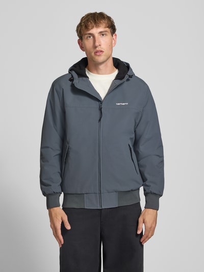 Männermodel trägt Funktionsjacke mit Logo-Stitching und Kapuze von Carhartt Work In Progress, grau, Reißverschluss, Ganzkörperansicht von vorne.