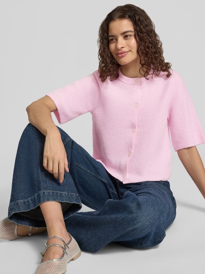 Frauenmodel trägt Boxy Fit Strickjacke aus Rippstrick Modell 'SILLY' von PIECES in Melange-Optik, kurzarm, Crew Neck, rosa Farbton, Material-Mix mit Woll-Anteil, einreihige Knopfleiste, Nahaufnahme Oberkörper.