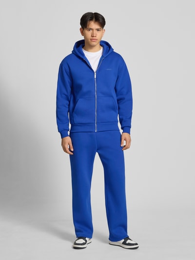 Männermodel trägt Sweatjacke mit Känguru-Tasche von REVIEW in Blau aus Baumwoll-Mix, Relaxed-Fit, mit Reißverschluss, Ganzkörperansicht von vorne