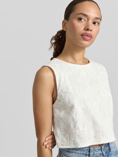 Frauenmodel trägt Cropped Blusentop mit rückseitigem Schlüsselloch-Ausschnitt von Mango aus Reine Baumwolle, Straight Fit, Nahaufnahme Oberkörper. Modell 'LISBOA', Rundhalsausschnitt, Rückenlänge bei Größe S: 42 cm. Strukturierte, floral gemusterte Oberfläche in Naturweiß.