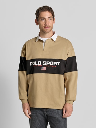 Mężczyzna nosi Relaxed Fit Poloshirt aus reiner Baumwolle od Polo Ralph Lauren w beżowym i czarnym kolorze, napis na piersi, Ganzkörperansicht von vorne.