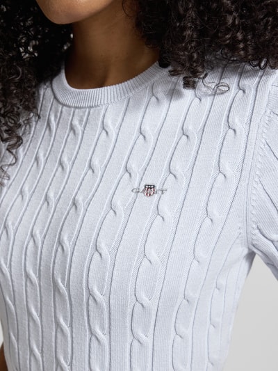 Kobiece modelka nosi Strickshirt mit Zopfmuster und Label-Stitching od Gant, mieszanka bawełny z dodatkiem streczu, krój slim fit, ścieg warkoczowy, wyhaftowane logo, okrągły dekolt, długość tyłu w rozmiarze S: 57 cm, długość rękawów w rozmiarze S: 24 cm, jasny odcień, detaliczny widok na górną część ciała