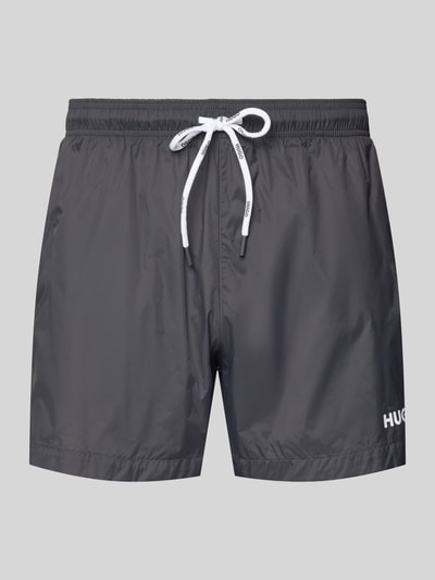 Produktfoto, Badeshorts mit Label-Print Modell 'HAITI' von HUGO w kolorze szarym, jednolita struktura, elastyczny pas, boczne kieszenie, kontekst: męskie kąpielówki, nadruk z logo, kieszeń z tyłu, tunel do wiązania