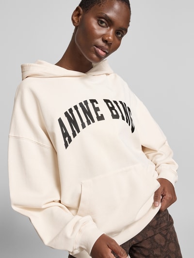 Frauenmodel trägt Oversized Hoodie mit Logo-Print von Anine Bing in Creme, Reine Baumwolle, Känguru-Tasche, elastische Abschlüsse, Oversized-Fit, Rückenlänge bei Größe S 68 cm, Nahaufnahme Oberkörper.