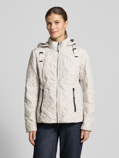 Modelka nosi Jacke mit Stehkragen und Reißverschlusstaschen od Gil Bret, jasny beż, pikowana bawełna, krój slim fit, kaptur, dwie kieszenie na suwak, ochrona przed pogodą, długość tyłu w rozmiarze 36: 61 cm, szerokość ramion w rozmiarze 36: 37 cm, szerokość w rozmiarze 36: 50 cm, długość rękawa w rozmiarze 36: 64 cm, widok całej sylwetki z przodu.