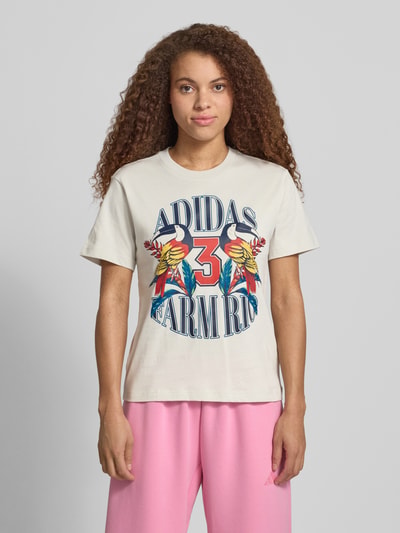 Vrouwenmodel draagt T-shirt mit Label-Print van adidas Sportswear in beige, puur katoen, regular fit, ronde hals, ruglengte maat S 61 cm, labelprint in kleurrijk design, voorkant, Ganzkörperansicht von vorne.