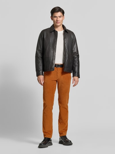 Männermodel trägt orangefarbene Straight Fit Cordhose aus Baumwoll-Mix Modell 'Mythos Flex Cord' von Brax, kombiniert mit dunkler Lederjacke, hellem Shirt und schwarzen Schuhen, schlichte Studioumgebung, Ganzkörperansicht von vorne, Herren Cordhose von Brax.
