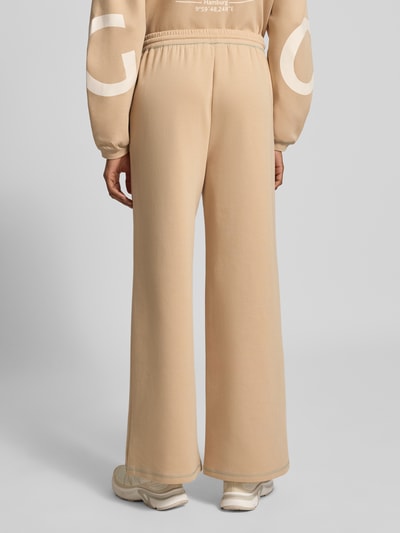 Frauenmodel trägt Wide Fit Sweatpants aus Baumwoll-Mix Modell 'Gunda' von GUIDO MARIA KRETSCHMER WOMAN in Beige, elastischer Bund, Eingrifftaschen, Logo-Print, glatte Textur, Ganzkörperansicht Rückansicht.