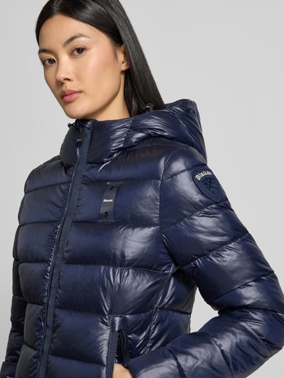 Frauenmodel trägt dunkelblaue Jacke in Stepp-Optik Modell 'CAROLL' von Blauer USA mit Logo-Detail und Kapuze, Nahaufnahme Oberkörper.