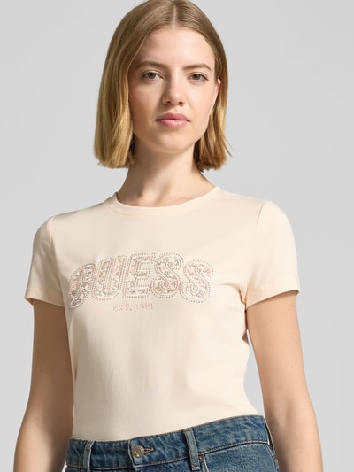 Frauenmodel trägt Slim Fit T-Shirt mit Strasssteinbesatz von Guess in Beige, Baumwoll-Mix, Rundhalsausschnitt, Logo-Detail, Nahaufnahme Oberkörper.