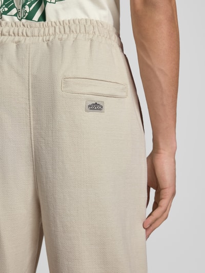 Männermodel trägt Track Pants mit Logo-Stitching Modell 'Skagen' von Pegador in Beige aus Reine Baumwolle mit Gesäßleistentasche, Nahaufnahme Rückansicht. Loose-Fit, elastischer Bund, französische Eingrifftaschen, Bundweite 40 cm, Innenbeinlänge 78 cm, Größe M.