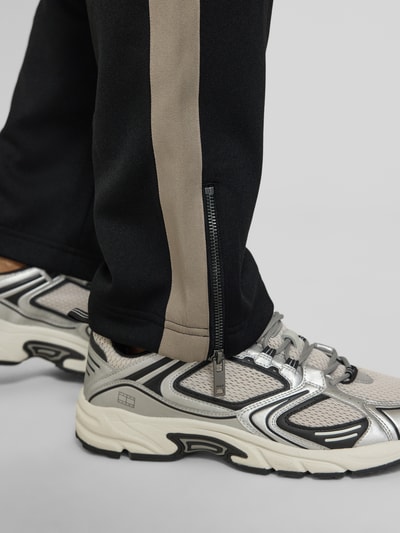 Männermodel trägt Regular Fit Jogpants mit Baumwoll-Anteil Modell 'DOLOS' von HUGO, schwarz mit beigem Kontraststreifen, Sweat-Textur, Reißverschluss am Bein, Label Print, Nahaufnahme Unterschenkel