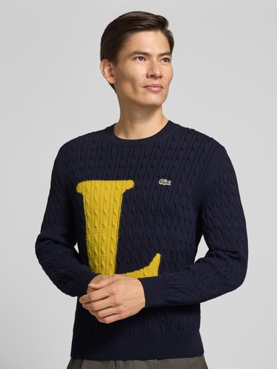 Männermodel trägt Regular Fit Strickpullover aus Baumwoll-Mix von Lacoste in Dunkelblau mit gelbem L, strukturierte Oberfläche, Nahaufnahme Oberkörper. Herren Strickjacke von Lacoste.