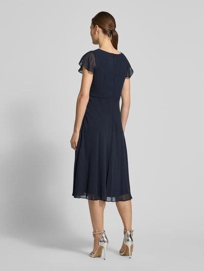 Frauenmodel trägt Knielanges Cocktailkleid mit Ziersteinbesatz von Christian Berg Cocktail in Dunkelblau, Chiffon, taillierter Schnitt, Rückansicht Ganzkörperansicht.