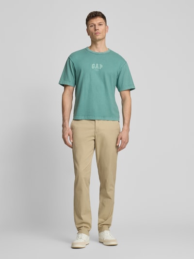 Männermodel trägt Slim Fit Chino mit Eingrifftaschen von GAP aus Baumwoll-Elasthan-Mix, beige Farbe, Ganzkörperansicht von vorne.