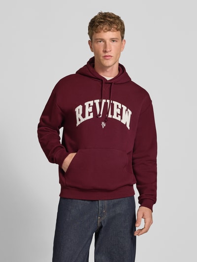 Męski model nosi Relaxed Fit Hoodie aus Baumwoll-Mix mit Logo-Stickerei von REVIEW w kolorze bordowym, wyhaftowane logo, kaptur z tunelem, kieszeń kangurka, krój relaxed fit, długość tyłu w rozmiarze M: 67 cm, długość rękawów w rozmiarze M: 65 cm, prążkowane wykończenia, widok całej sylwetki z przodu.