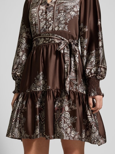 Frauenmodel trägt sukienkę do kolan z dekoltem w serek, model 'MALFIDA' od Neo Noir, taliowany krój, pasek w talii, listwa guzikowa, długość tyłu 92 cm, szerokość klatki piersiowej 43 cm, długość rękawów 64 cm, brązowy wzór, połyskliwa tekstura, detaliczna faktura, nahaufnahme okolicy talii.