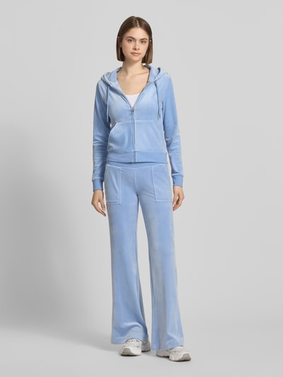 Frauenmodel trägt Slim Fit Sweatjacke aus Samt Modell 'Robertson' von Juicy Couture im hellblauem Elastischer-Material-Mix mit Kapuze, Reißverschluss, geteilte Känguru-Tasche und elastische Abschlüsse, Ganzkörperansicht von vorne.