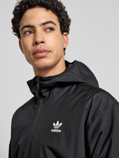 Mężczyzna nosi Jacke mit Kapuze od adidas Originals, czarny kolor, logo, krój regular fit, kaptur, zamek, zbliżenie na górną część ciała. Długość pleców w rozmiarze M 71 cm, szerokość ramion w rozmiarze M 55 cm, szerokość klatki piersiowej w rozmiarze M 61 cm, długość rękawa w rozmiarze M 66 cm.